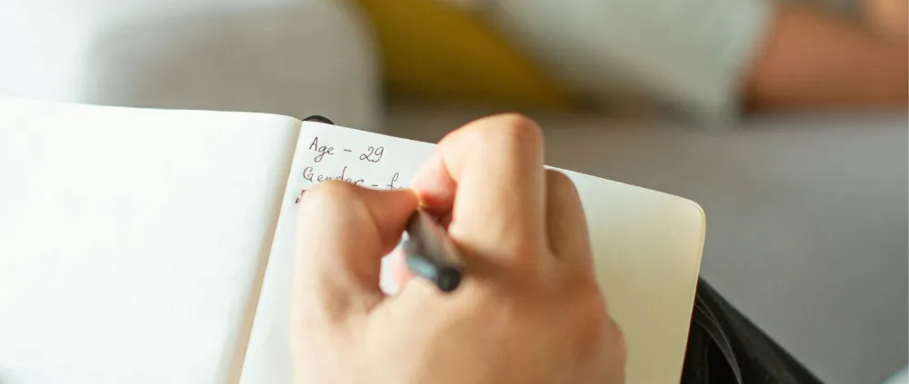 Main d'une personne qui prend des notes dans un carnet, une personne allongée sur un canapé à l'arrière-plan