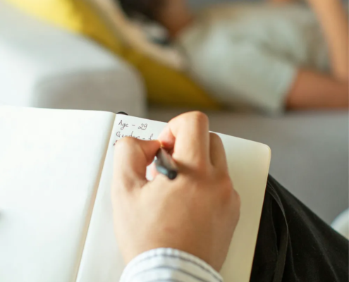 Main d'une personne qui prend des notes dans un carnet, une personne allongée sur un canapé à l'arrière-plan