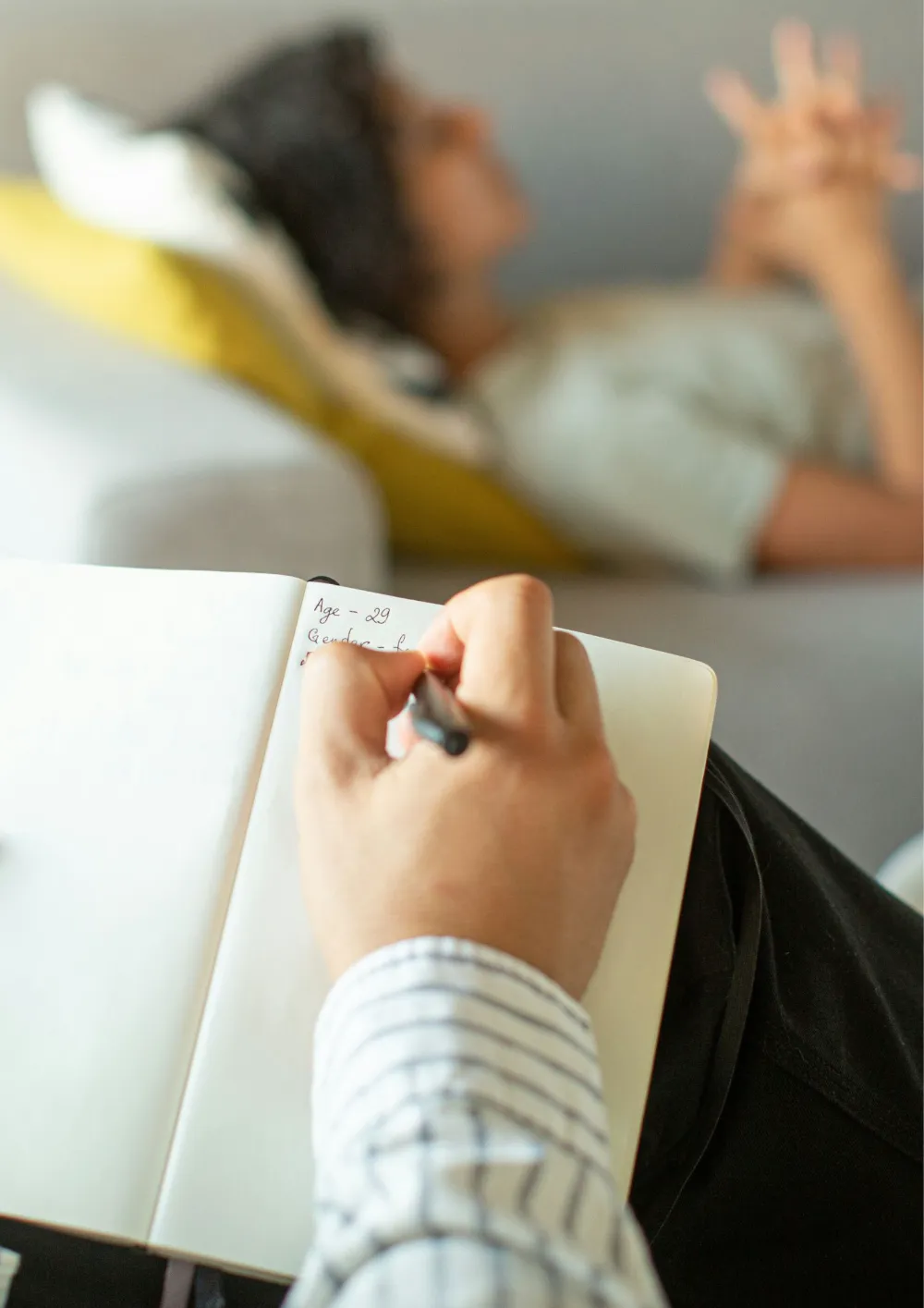 Main d'une personne qui prend des notes dans un carnet, une personne allongée sur un canapé à l'arrière-plan